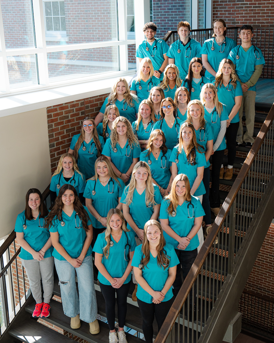A group of nursing students wearing scrubs and stethoscopes stand together on a stairwell inside an academic building during a cohort photo.